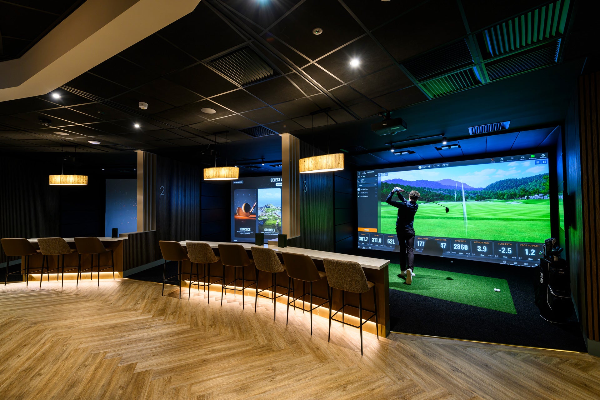 Golf simulator room with large screen displaying a golf game, surrounded by chairs.